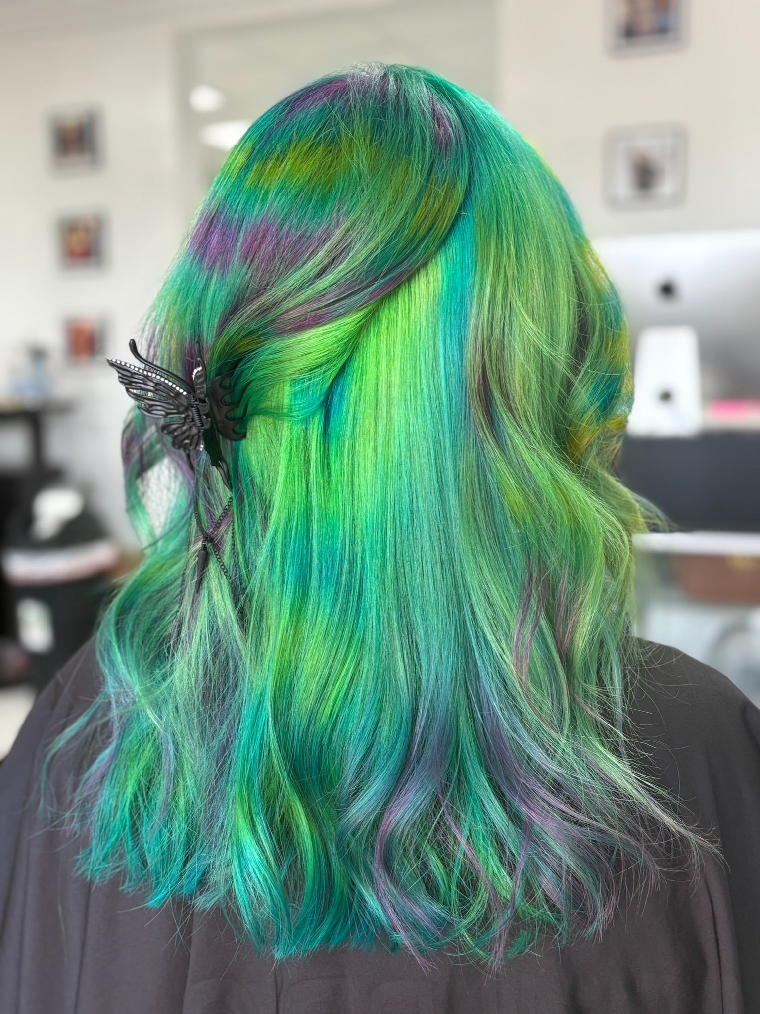 Person with vibrant green and blue hair styled in loose waves, wearing a black butterfly hair accessory.